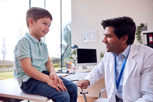 Male Doctor Or GP Wearing White Coat Examining Smiling Boy Testi
